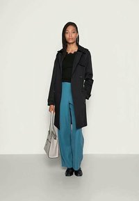 Schwarzer Trenchcoat mit einem doppelbrüstigen Design, kombiniert mit türkisfarbenen weit geschnittenen Hosen und einer grauen Handtasche. Glatter Stoff mit strukturiertem Schnitt.