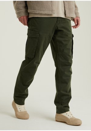 Personne portant un pantalon cargo vert olive, une veste polaire beige et des baskets beiges, debout devant un fond clair uni.