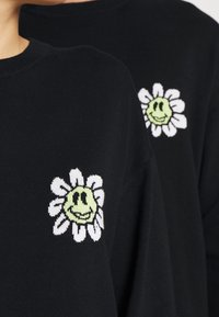 Maglione nero con un design di margherita bianca e un viso verde sorridente. Texture morbida, scollo a girocollo classico e polsini a coste.