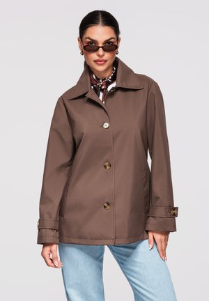 Femme portant une veste marron à boutons, un jean bleu clair, de petites lunettes de soleil foncées et un foulard à motifs autour du cou, sur un fond uni.