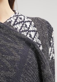 Suéter de punto gris oscuro con un aspecto texturizado, con un patrón geométrico blanco en los hombros. Material suave.