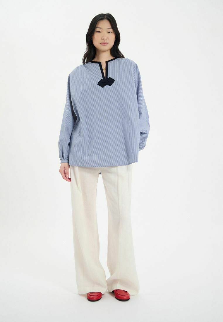 Chemise rayée bleu et blanc avec une patte de boutonnage et un détail en forme de cœur noir, assortie à un pantalon large blanc et des chaussures rouges.