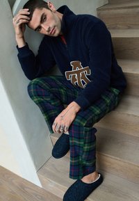 Marineblauer Fleecepullover mit einem beigefarbenen Logo, kombiniert mit grün-blau karierten Hosen und dunklen Hausschuhen zum Hineinschlüpfen. Im Hintergrund sind Holzstufen zu sehen.