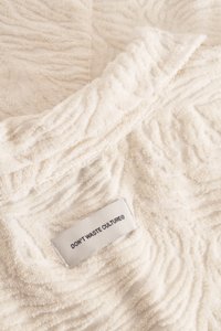 Crèmekleurige handdoek met een gestructureerd, golvend patroon. Heeft een label met de tekst "VERSPIL DE CULTUUR®," vastgezet met ton sur ton stiksels.