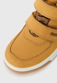 Chaussure en suède tan avec trois sangles Velcro, bout arrondi, semelle blanche contrastante et détails texturés. Présente des éléments de marque sur le côté.