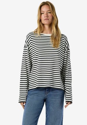 Jeune femme aux longs cheveux blonds portant une chemise ample à manches longues rayée noir et blanc et un jean bleu, debout devant un fond blanc.