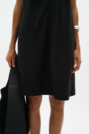 Donna che indossa un vestito nero fino al ginocchio, tiene una borsa nera nella mano sinistra e indossa un braccialetto bianco al polso destro.