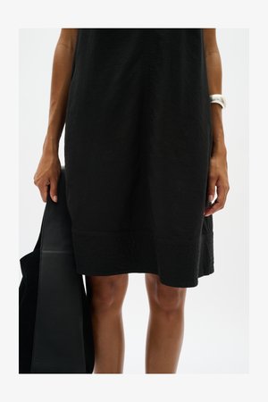 Donna che indossa un vestito nero fino al ginocchio, tiene una borsa nera nella mano sinistra e indossa un braccialetto bianco al polso destro.