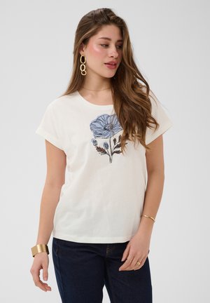 Kvinde med langt brunt hår iført en hvid t-shirt med et blåt blomsterprint, guldbøjler, armbånd og mørke jeans, der kigger til siden.