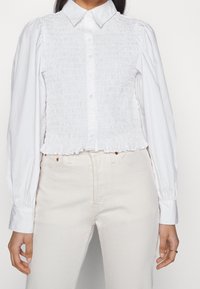 Hvid bluse med smocket liv og lange puffede ærmer. Skjorten har en knaplukning fortil og en kort, scallopet kant. Sammen med lyse jeans.