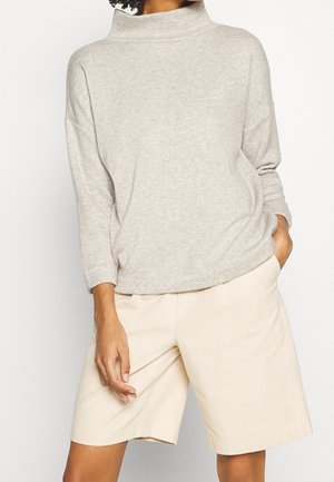 Personne portant un haut gris clair à manches longues avec un col montant et un short ample beige longueur genou, une main dans la poche.