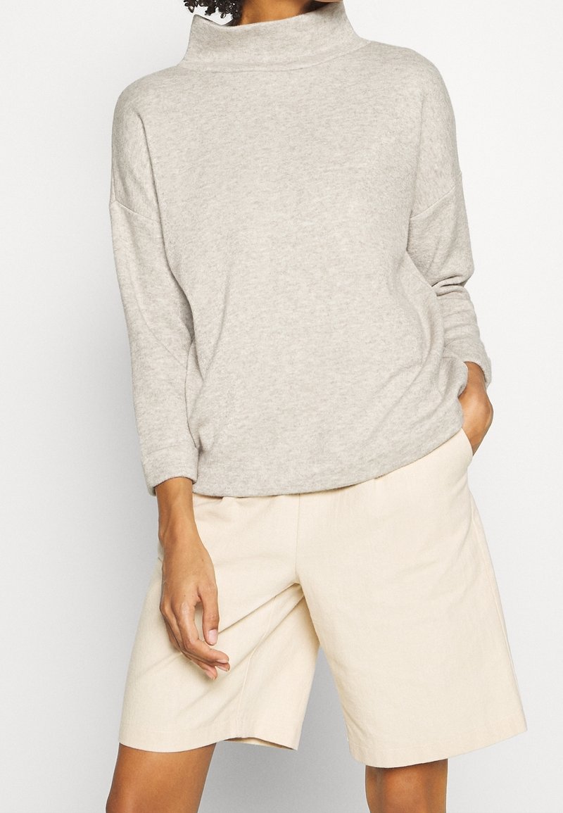 Personne portant un haut gris clair à manches longues avec un col montant et un short ample beige longueur genou, une main dans la poche.