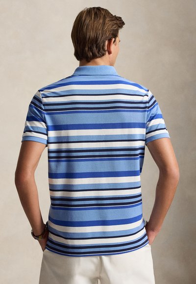 Homme portant un polo à manches courtes à rayures horizontales bleu, blanc et noir, et un short blanc, de dos avec les mains dans les poches.