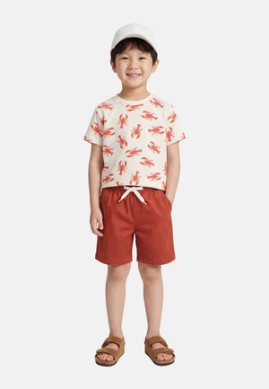 Enfant souriant portant une casquette blanche, un t-shirt crème à motif homard, un short rouge rouille avec un cordon blanc, et des sandales marron, debout les mains dans les poches.
