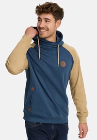 Sudadera con capucha que presenta un cuerpo azul marino y mangas raglán beige. Incluye un cordón, parche con el logo y detalles de cuero en los bolsillos.
