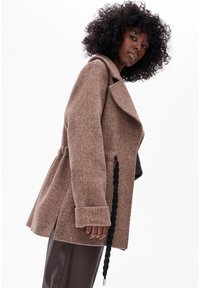 Bruine wolmix jas met een gelapt kraag, oversized ontwerp en een zwarte trekkoorddetail aan de zijkant. Textiel met structuur en lange mouwen.