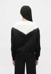 Czarna bluza z białym wzorem w kształcie szewrona na górnej części pleców, ozdobiona frędzlami. Długie rękawy i luźny krój. Teksturowany materiał.