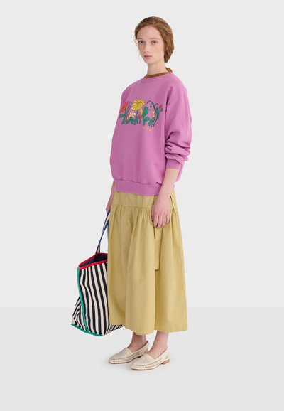 Jeune femme portant un sweat-shirt rose à motifs floraux et une jupe midi beige, tenant un sac fourre-tout rayé, debout devant un fond uni.