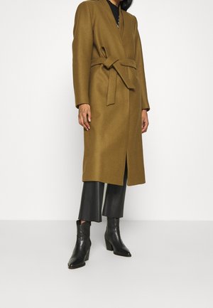 Personne portant un long manteau marron ceinturé, un pantalon noir court et des bottines noires à talons, se tenant devant un fond blanc uni.