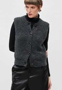 Halline hall villane varrukateta vest, millel on tekstuuriga viimistlus, nupuga kinnitamine ja kergelt ümar hem. Kandmine musta kõrge kaelusega särk üle.