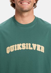 Žalia trumpa rankovė t-marškiniai pagaminti iš medvilnės, su išlenktu geltonos ir baltos spalvos "QUIKSILVER" logotipu per krūtinę. Minkšta tekstūra, klasikinis siluetas.