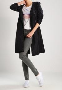 Černý dlouhý trenčkot s kapucí, nošený přes bílý tričko s grafikou, v kombinaci se slim šedými kalhotami a bílými teniskami. Hladký matný povrch.