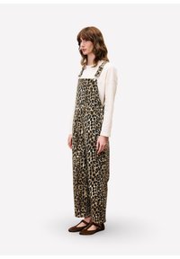 Vrouw in een beige langslievend shirt, met een luipaardprint overall en bruine Mary Jane-schoenen, staande tegen een eenvoudige lichte achtergrond.