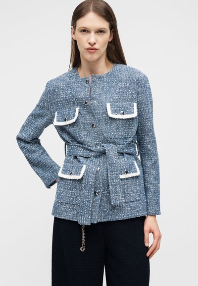 Γυναίκα που φοράει μπλε τουίντ σακάκι με ζώνη, ασημένια κουμπιά και τσέπες με καπάκι και λευκή επένδυση, συνδυασμένο με μαύρο παντελόνι.