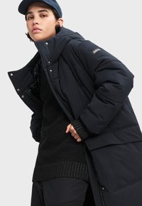 Námornícka puffová bunda s vysokým golierom, so zipsom a sponkami. Pod ňou je štruktúrovaný čierny sveter. Hladká tkanina s vypolstrovanými sekciami.