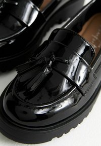 Czarne lakierowane loafersy z grubą czarną gumową podeszwą, dekoracyjnym detalem w postaci frędzli oraz gładką, błyszczącą powierzchnią.