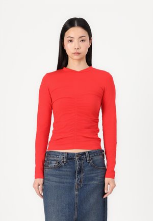 Maglietta rossa a maniche lunghe con un design aderente e una texture arricciata sul davanti, abbinata a una gonna di denim. Tessuto morbido ed elastico.