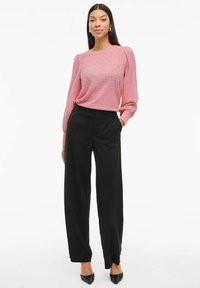 Top lungo a maniche lunghe di un rosa chiaro strutturato con spalle a palloncino, abbinato a pantaloni neri a vita alta a gamba larga e scarpe nere a punta.