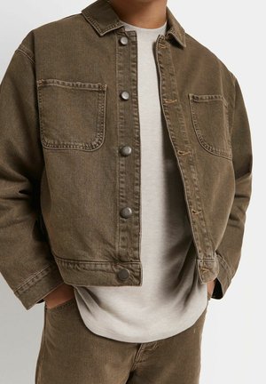 Uomo che indossa una giacca di jeans marrone con bottoni a pressione sopra una maglietta beige chiaro e pantaloni marroni abbinati, mani in tasca, sfondo neutro.