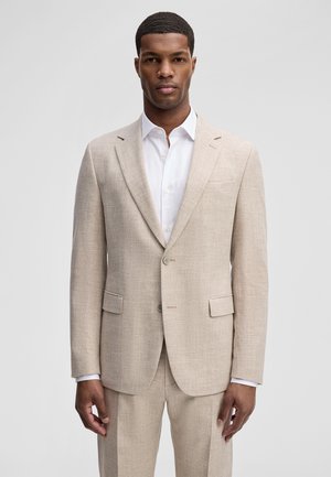 Mężczyzna ubrany w jasnobeżową marynarkę i dopasowane spodnie, na białej koszuli, stojący na jednolitym tle.