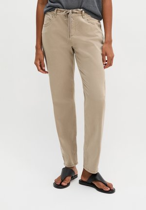 Personne portant un pantalon beige à cordon, une chemise grise et des sandales noires ouvertes, debout devant un fond clair uni.