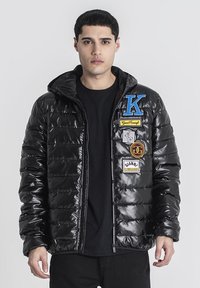 Chaqueta acolchada negra con un acabado brillante, que presenta costuras horizontales, una capucha y varios parches coloridos en el frente.