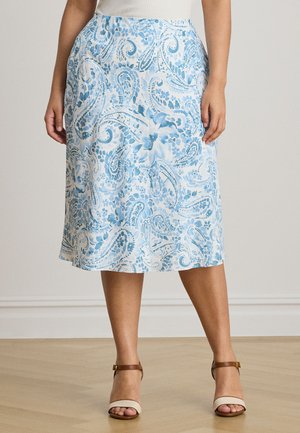 Žena v bielej sukni po kolená s modrým kvetinovým paisley vzorom a hnedými sandálmi s remienkom okolo členka, stojaca na drevenej podlahe.