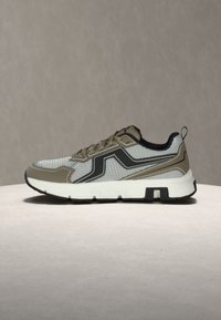Baskets de sport gris clair et beige avec accents noirs, tige en mesh, semelle blanche et languette au talon, présentées sur une surface neutre.