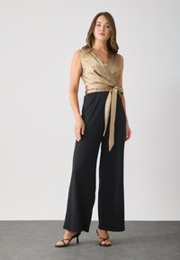 Saténový overal s taupe tělem, černými širokými kalhotami a detailem zavázaného pasu. Černé sandály na vysokém podpatku doplňují outfit.