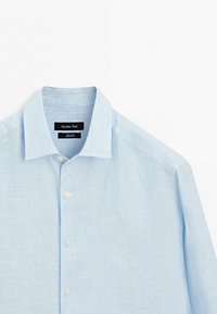Chemise bleu clair à manches longues avec boutons, fines rayures blanches et col spread, posée à plat sur une surface blanche.