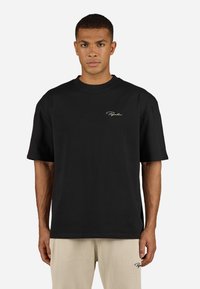 Mann iført svart oversized T-skjorte med lite hvitt logo på brystet og beige rillede bukser med logo på venstre lår, stående mot hvit bakgrunn.