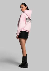 Hoodie oversize rosa con testo nero sulla schiena, abbinato a una minigonna nera e stivali alti fino al ginocchio neri. Tessuto morbido, vestibilità rilassata.