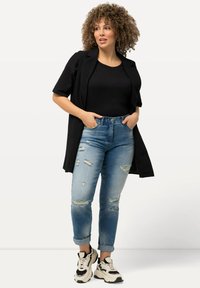 Blazer negro de manga corta sobre una blusa negra, combinado con jeans desgastados de un azul claro y zapatillas gruesas en blanco y negro.