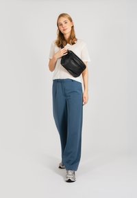 Fekete bőrcrossbody táska, fehér könnyű póló, kék bővebb nadrág és ezüst sneaker. A háttér egyszínű fehér.