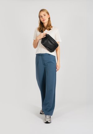Borsa a tracolla nera in pelle, t-shirt bianca leggera, pantaloni larghi blu e sneakers argento. Sullo sfondo c'è un bianco uniforme.