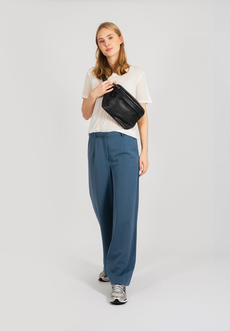 Fekete bőrcrossbody táska, fehér könnyű póló, kék bővebb nadrág és ezüst sneaker. A háttér egyszínű fehér.