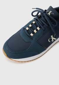 Marineblauwe atletische sneaker van suede en nylon, met crèmekleurige veters, witte middenzool en gouden CK-logo aan de zijkant. Geprofileerde rubberen buitenzool.