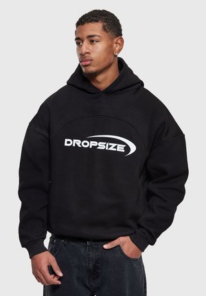 Ung mand iført en sort hættetrøje med "DROPSIZE"-logo, der kigger til siden, med den ene hånd i lommen, mod en ensartet lys grå baggrund.