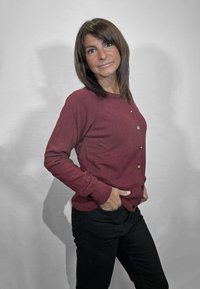 Cardigan color marrone scuro con chiusura a bottoni sul davanti, polsini e orlo a coste, e vestibilità rilassata, abbinato a pantaloni neri. Tessuto liscio, design minimale.