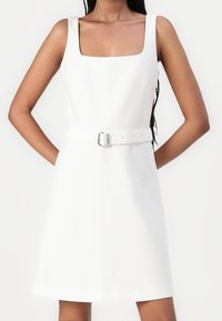 Vit klänning med fyrkantig halsringning, breda axelremmar, figursydd midja och ett matchande spännbälte. Slät struktur med en utsvängd fåll.